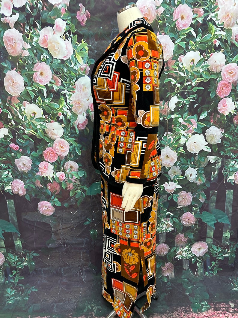 May include: A black velvet skirt suit with a geometric and floral print in orange, brown, and white. The suit features a long skirt with a side slit and a matching jacket with a collar.