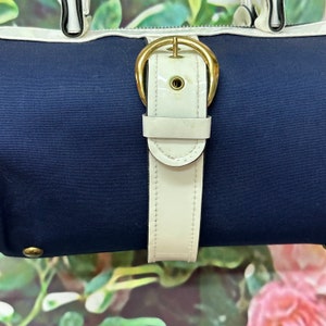 May include: A navy blue canvas bag with a white leather strap and gold buckle. The bag has a white trim around the top.