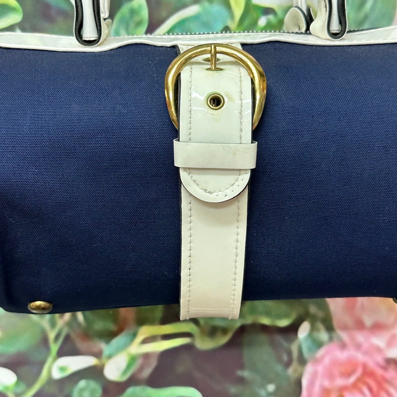 May include: A navy blue canvas bag with a white leather strap and gold buckle. The bag has a white trim around the top.