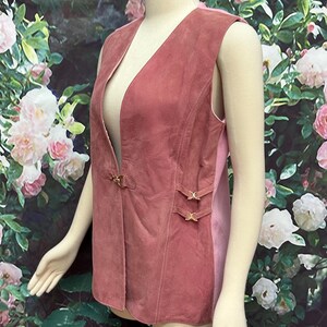 May include: A sleeveless suede vest in a dusty rose color with gold buckle accents. The vest has a deep V-neck and a slightly fitted silhouette.