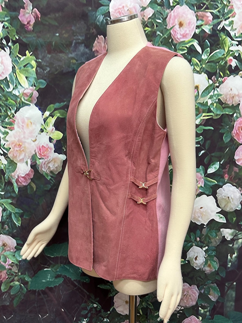 May include: A sleeveless suede vest in a dusty rose color with gold buckle accents. The vest has a deep V-neck and a slightly fitted silhouette.