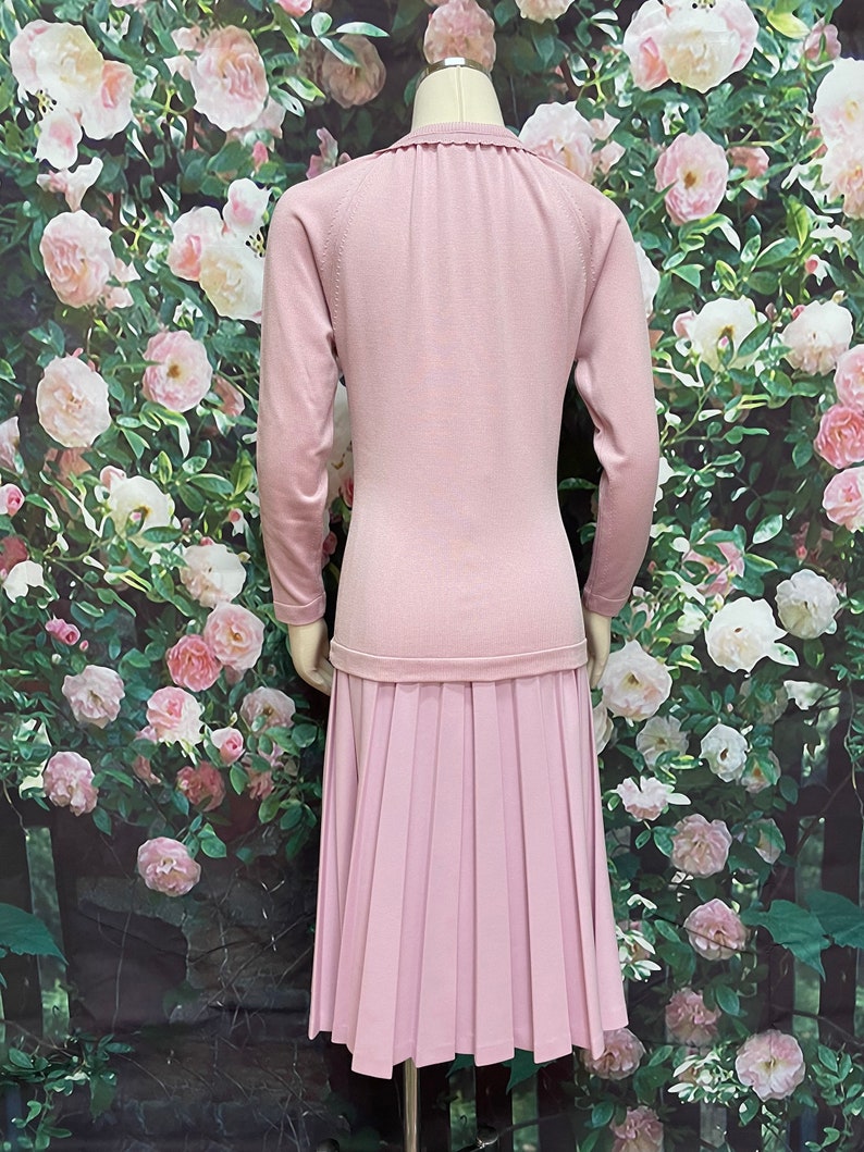 May include: A light pink knitted dress with a pleated skirt. The dress has a round neckline and long sleeves.