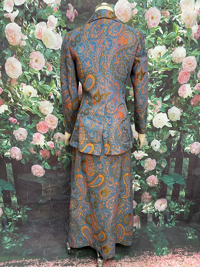 May include: A blue and orange paisley patterned skirt suit. The jacket has a notched collar and long sleeves. The skirt is floor-length.