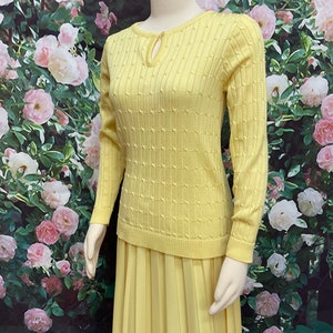 May include: A yellow knitted sweater with a keyhole neckline and a matching pleated skirt. The sweater has a textured pattern and long sleeves.