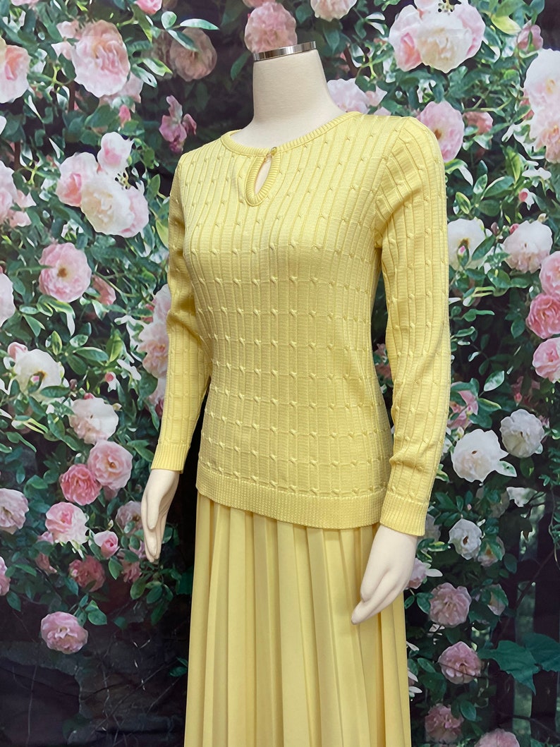 May include: A yellow knitted sweater with a keyhole neckline and a matching pleated skirt. The sweater has a textured pattern and long sleeves.