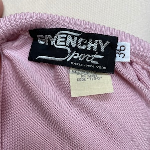 May include: A pink knitted sweater with a black Givenchy Sport label. The label has the text "Givenchy Sport" in white lettering and "Paris - New York" in smaller white lettering. The label also has a white tag with the text "Separate - Machine Wash - RN 34692 - Code "T/G-Q"".