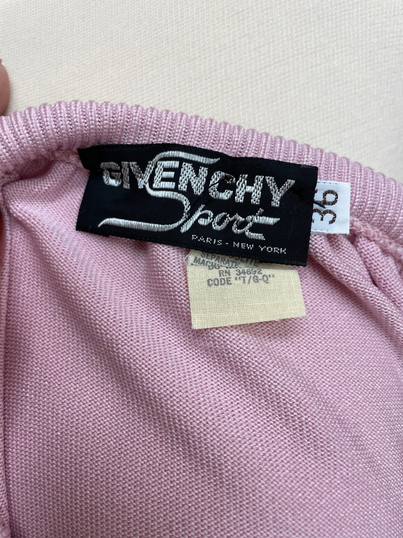 May include: A pink knitted sweater with a black Givenchy Sport label. The label has the text "Givenchy Sport" in white lettering and "Paris - New York" in smaller white lettering. The label also has a white tag with the text "Separate - Machine Wash - RN 34692 - Code "T/G-Q"".