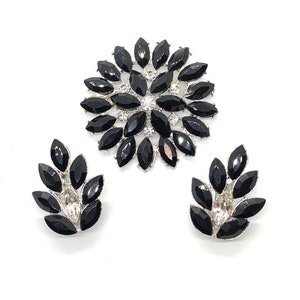 May include: A black and silver rhinestone brooch and matching earrings. The brooch is a large flower shape with black rhinestones and a centre of clear rhinestones. The earrings are smaller and have a similar design.