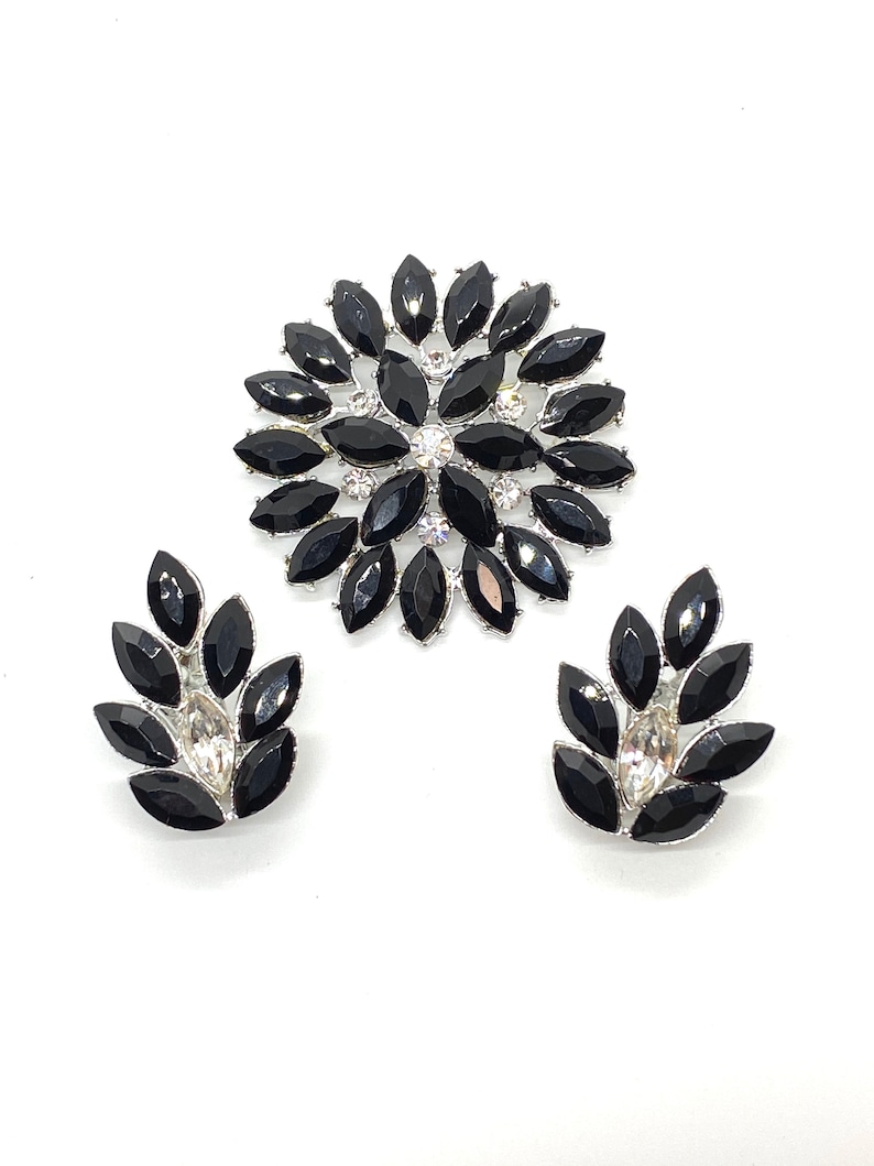 May include: A black and silver rhinestone brooch and matching earrings. The brooch is a large flower shape with black rhinestones and a centre of clear rhinestones. The earrings are smaller and have a similar design.