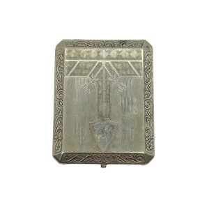 May include: A silver cigarette case with an ornate design. The case has a raised shield in the center and a decorative border.