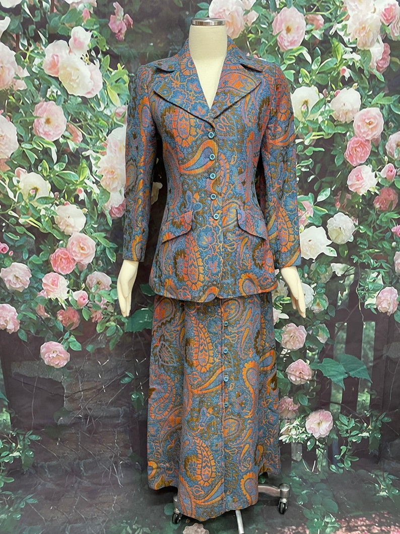 May include: A blue and orange paisley print skirt suit with a fitted jacket and a long skirt. The suit is on a mannequin against a backdrop of pink roses.