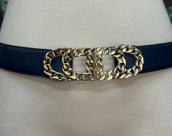 May include: A navy blue belt with a gold-colored chain-link buckle. The buckle is a double-loop design with a central bar. The belt is displayed on a mannequin.