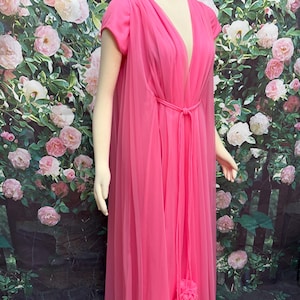 May include: A floor-length hot pink gown featuring a deep V-neckline, short sleeves, and a sheer layered overlay. The garment includes a thin matching tie belt at the waist and two decorative fabric pom-poms hanging from the center front.