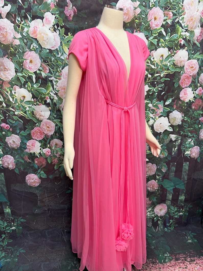 May include: A floor-length hot pink gown featuring a deep V-neckline, short sleeves, and a sheer layered overlay. The garment includes a thin matching tie belt at the waist and two decorative fabric pom-poms hanging from the center front.