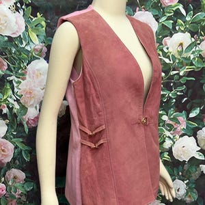 May include: A sleeveless suede vest in a dusty rose color with a deep V-neckline and a single button closure. The vest has a fitted silhouette and features decorative stitching and a small buckle detail on the side.