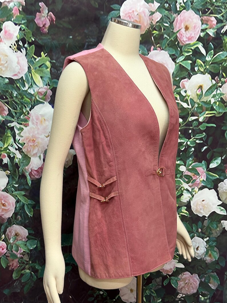 May include: A sleeveless suede vest in a dusty rose color with a deep V-neckline and a single button closure. The vest has a fitted silhouette and features decorative stitching and a small buckle detail on the side.