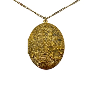May include: A gold-toned oval locket necklace with a detailed embossed design of a village scene. The locket features a tree, houses, and figures.