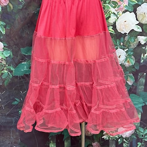 May include: A bright pink petticoat with a layered design. The top section is solid, while the lower portion features multiple tiers of sheer, ruffled fabric. The petticoat is displayed on a metal stand against a floral backdrop.