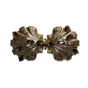 May include: A gold-toned metal clasp with a symmetrical design. The clasp features a fan-like shape with ridged detailing and is adorned with several dark gray, oval-shaped embellishments. The clasp is designed to connect two pieces.