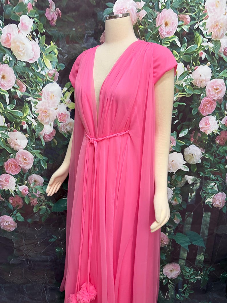 May include: A floor-length hot pink gown featuring a deep V-neckline, short cap sleeves, and a sheer chiffon overlay. The dress includes a matching thin fabric tie belt at the waist that creates vertical pleats and a flowing silhouette.