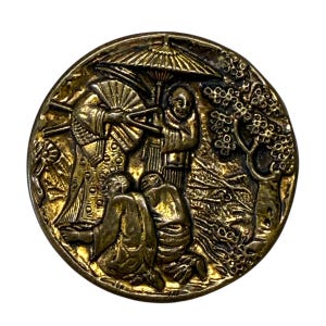 May include: A circular antique brass button with a detailed relief scene. The design features figures under a parasol and a tree, with intricate details. The button has a vintage aesthetic and a slightly tarnished finish.