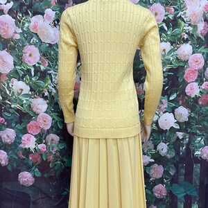 May include: A yellow knitted sweater and matching pleated skirt set. The sweater has a cable knit pattern and long sleeves. The skirt is knee-length and has a smooth texture.