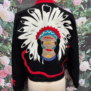 May include: A black jacket featuring a large, detailed appliqué on the back depicting a person wearing a white feathered headdress with red, blue, and yellow accents. The jacket has a red collar and a red curved line detail along the lower back.