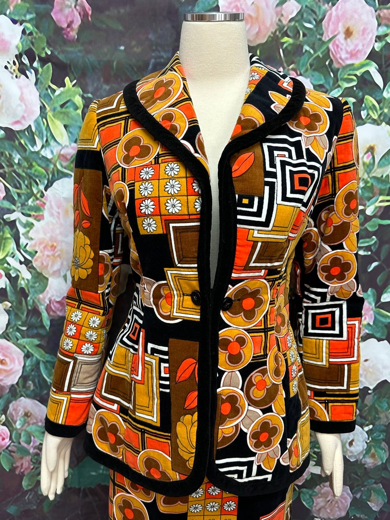 May include: A black, orange, and brown patterned jacket with a black velvet collar and trim. The jacket has a button closure and a fitted silhouette.