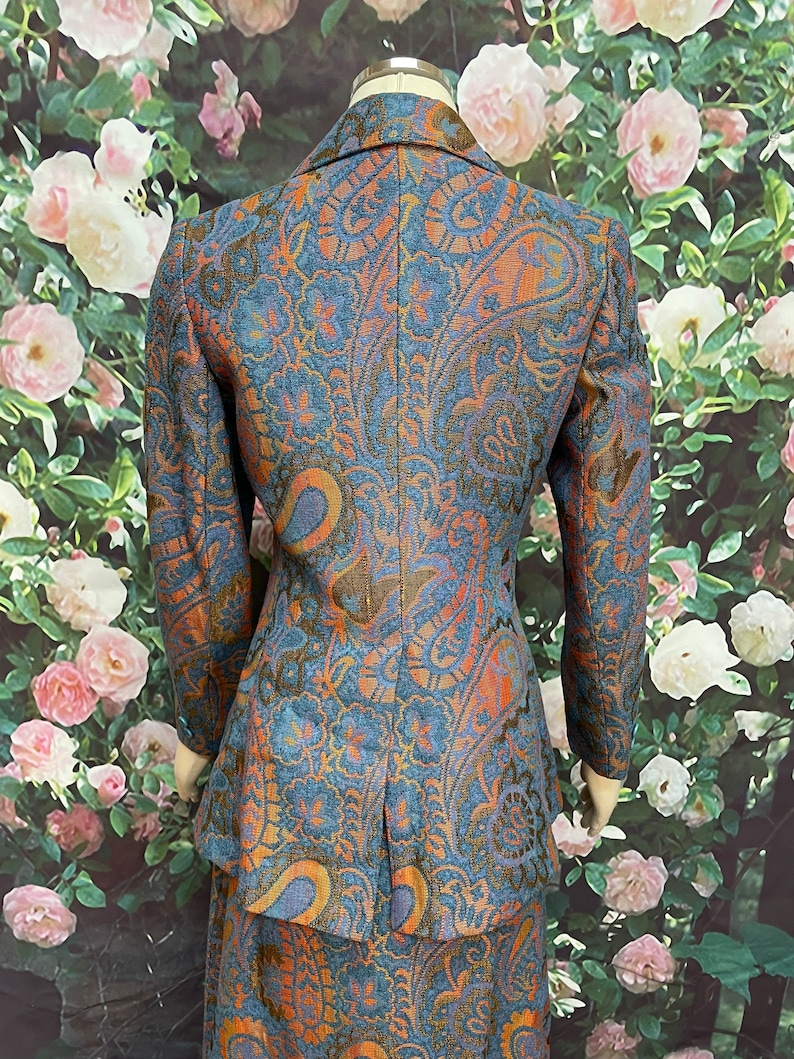 May include: A blue, orange and gold paisley patterned blazer and skirt set. The blazer has a notched collar and long sleeves. The skirt is A-line and falls to the knee.