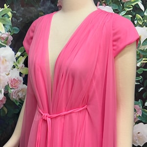 May include: A bright pink dress featuring a deep V-neckline and short sleeves. The garment is crafted with a sheer, layered tulle overlay that drapes over the bodice and skirt, secured at the waist with a matching thin pink fabric tie belt.