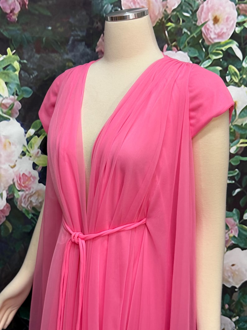 May include: A bright pink dress featuring a deep V-neckline and short sleeves. The garment is crafted with a sheer, layered tulle overlay that drapes over the bodice and skirt, secured at the waist with a matching thin pink fabric tie belt.
