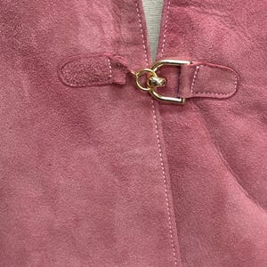 May include: A close-up of a pink suede jacket with a gold metal buckle closure.