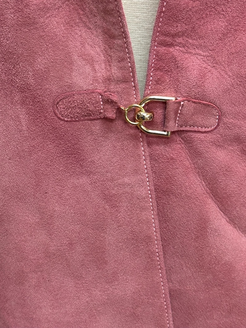 May include: A close-up of a pink suede jacket with a gold metal buckle closure.