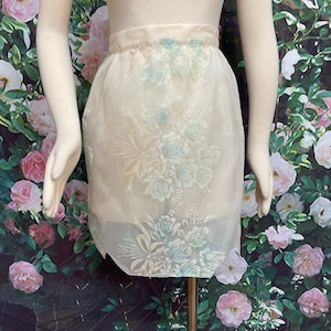 May include: A white sheer apron with a floral pattern in shades of green and blue. The apron has a gathered waist and a scalloped hem.