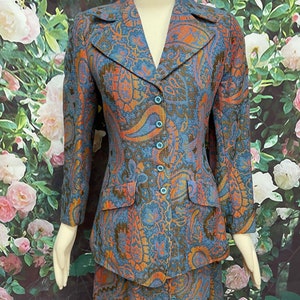 May include: A blue, orange and gold paisley patterned skirt suit. The jacket has a notched collar and a button closure. The skirt is knee-length.