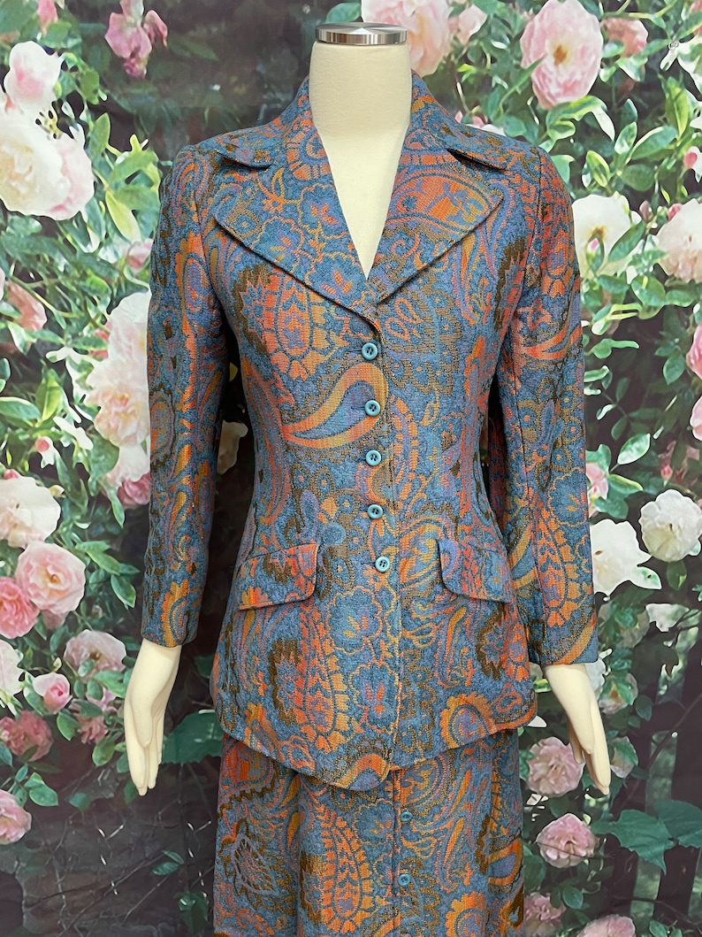 May include: A blue, orange and gold paisley patterned skirt suit. The jacket has a notched collar and a button closure. The skirt is knee-length.