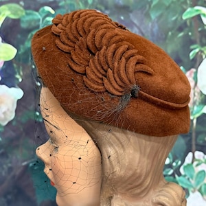 May include: A brown felt hat featuring a structured, rounded crown adorned with a series of layered, crescent-shaped felt appliques arranged in a decorative pattern. A delicate black netting veil with small, evenly spaced accents covers the front.