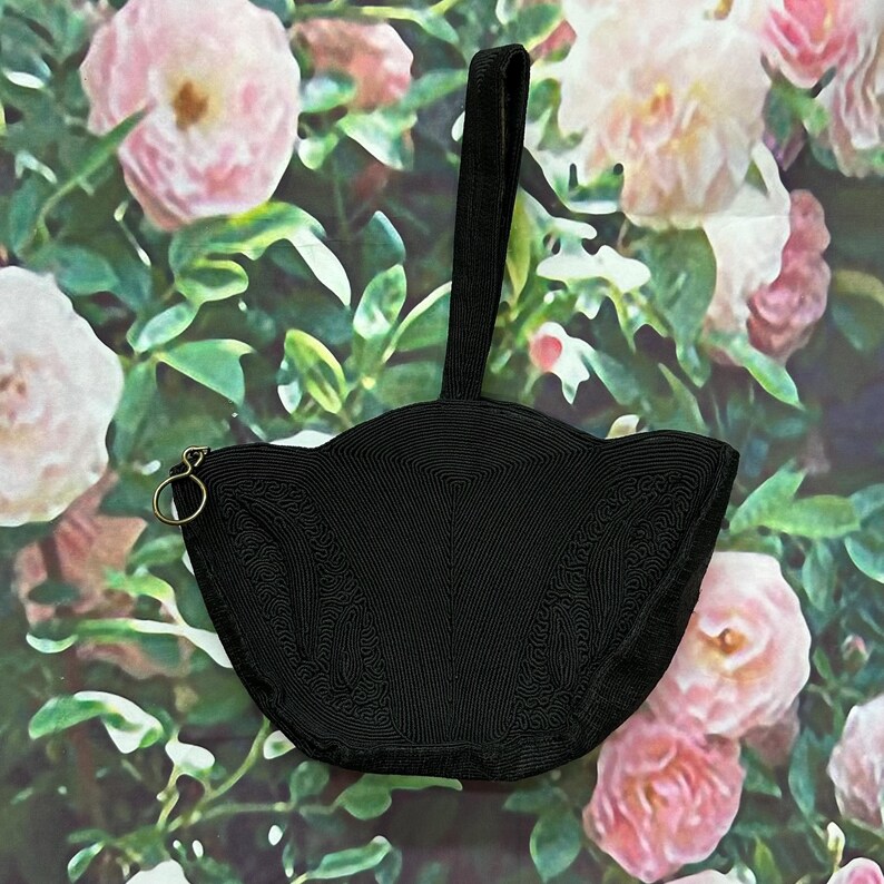 May include: A black beaded handbag with a scalloped shape and a single top handle. The bag is photographed against a backdrop of pink roses.