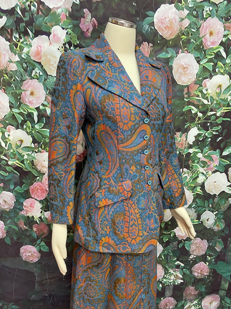 May include: A blue and orange paisley patterned skirt suit. The jacket has a notched collar and a single button closure. The skirt is knee-length.