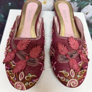 May include: A pair of burgundy slip-on shoes with intricate floral embroidery and tassel accents. The shoes feature a gold insole and the brand name "Amanda" is visible. The design includes pink, gold, and green embellishments.