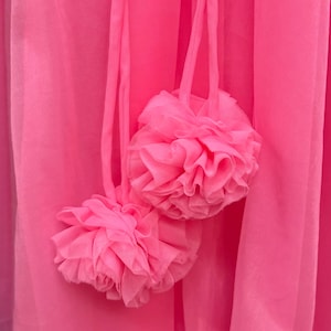 May include: A bright hot pink fabric garment featuring two decorative pom-poms made of layered, ruffled sheer material. The pom-poms are attached to the main fabric by thin, matching pink fabric cords, creating a textured and layered appearance.