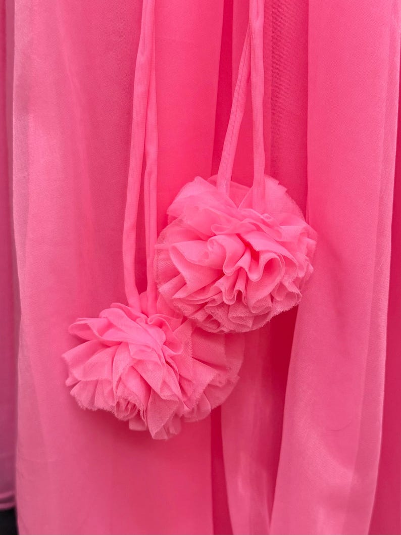 May include: A bright hot pink fabric garment featuring two decorative pom-poms made of layered, ruffled sheer material. The pom-poms are attached to the main fabric by thin, matching pink fabric cords, creating a textured and layered appearance.