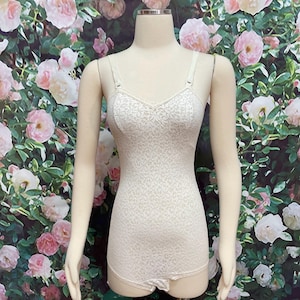 May include: A white lace bodysuit with a sweetheart neckline and adjustable straps. The bodysuit is on a white mannequin against a background of pink roses.