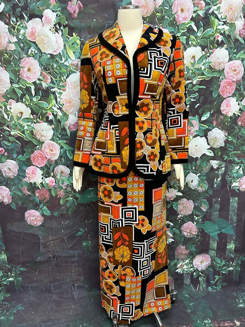 May include: A vintage 1970s style skirt suit in a bold geometric print with orange, brown, and black colors. The jacket has a black velvet trim and a single button closure. The skirt is a-line and falls to the knees.