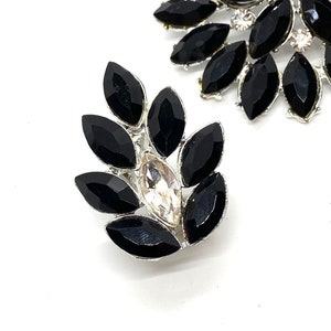May include: A pair of black and silver rhinestone brooches. The brooches are shaped like a flower with a large clear rhinestone in the centre.