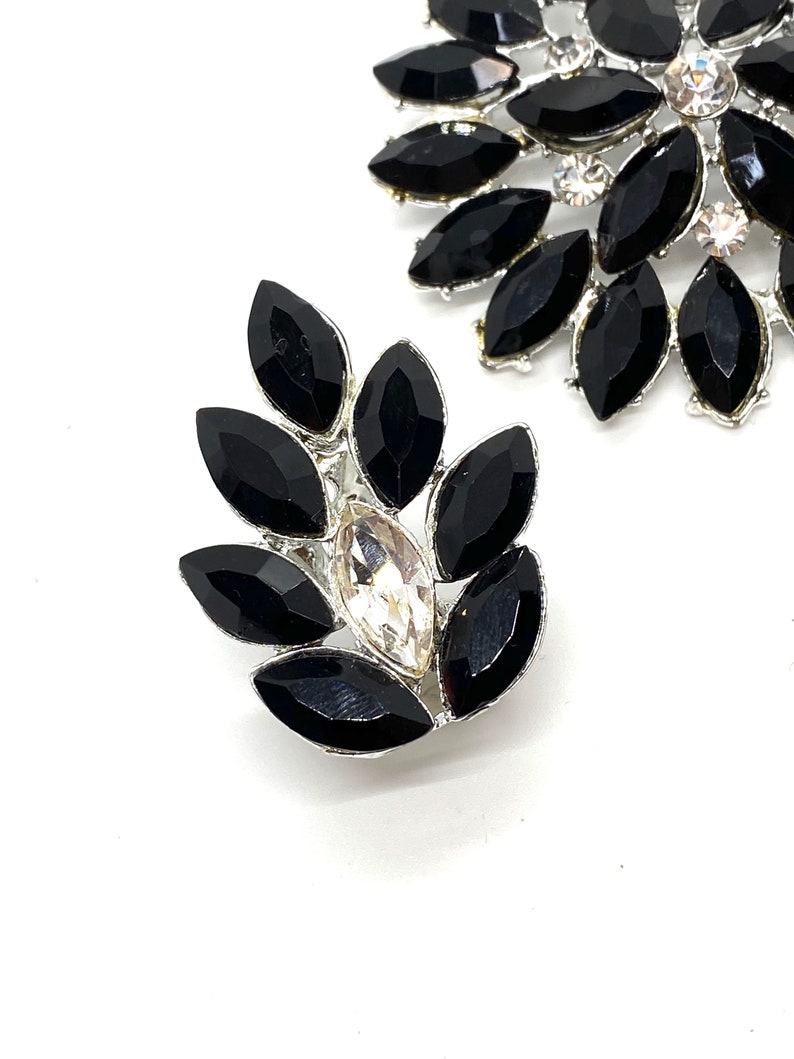 May include: A pair of black and silver rhinestone brooches. The brooches are shaped like a flower with a large clear rhinestone in the centre.