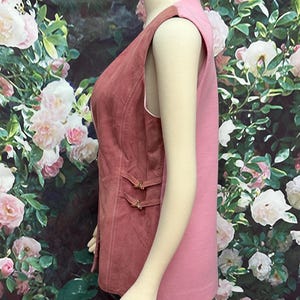May include: A sleeveless vest with a pink and rose-colored design. The vest has a button closure and a decorative strap detail on the front.