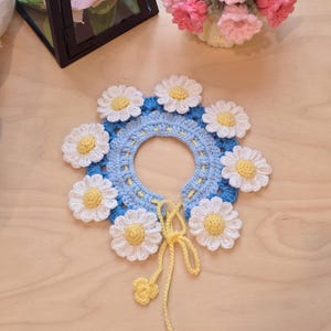 May include: A handmade crochet collar featuring a blue base adorned with white daisies with yellow centers. A yellow tie and three small flower accents hang from the collar. The collar is displayed on a light wooden surface.