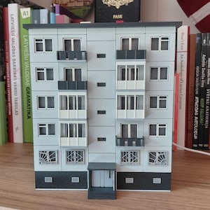 Può includere: Modello in miniatura di un condominio a più piani. L'edificio è dipinto di grigio chiaro con accenti grigio scuro sui balconi e sul tetto. Le finestre sono bianche e il modello è su una superficie di legno.