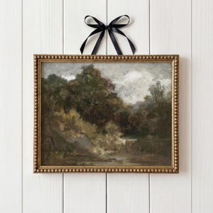 Pode incluir: Pintura de paisagem emoldurada com uma moldura dourada com contas, retratando uma cena de floresta com um rio. A pintura é pendurada com uma fita de cetim preta amarrada em um laço contra um fundo de madeira branca. A obra apresenta tons de verde, marrom e cinza.